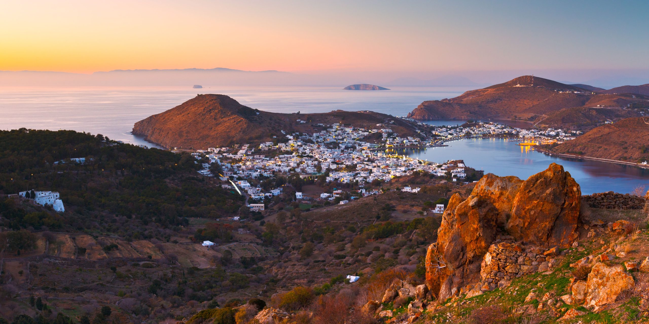 A sunset view of Patmos with a bay surrounded by hills and a town of whitewashed buildings. The calm sea reflects the soft evening light, and the rugged terrain adds depth to the landscape.