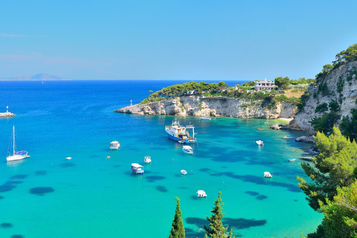 A clear bay in Alonissos with vibrant turquoise waters, small anchored boats, and a larger vessel in the center. The coastline features rocky cliffs, lush greenery, and a white building nestled among trees.