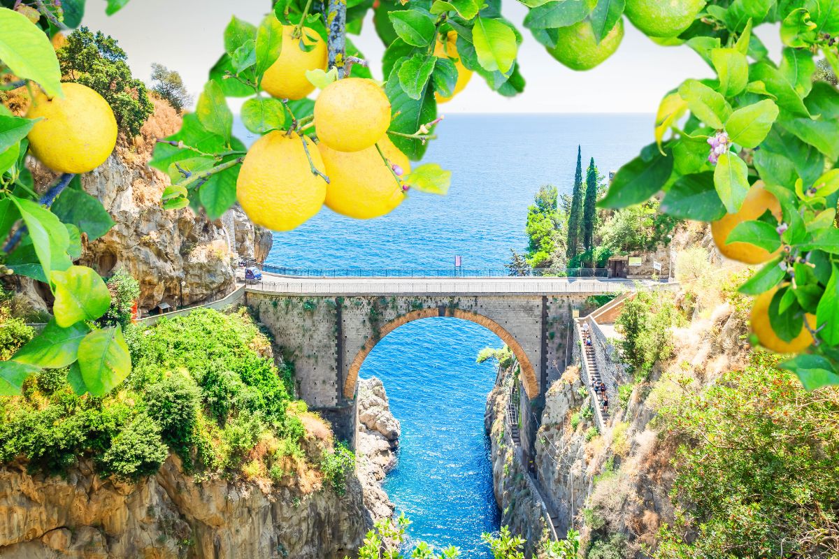 A stone bridge on the Amalfi Coast, Italy, spanning a narrow gorge with blue sea in the background. Yellow lemons hang from tree branches in the foreground_Weather in Italy and Greece in October