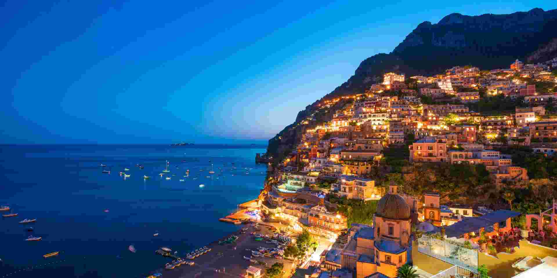 A stunning evening view of the Amalfi Coast, with hillside buildings illuminated against the deep blue sea and sky, and boats scattered along the shoreline. Amalfi Coast_planning a trip to italy and greece_feature