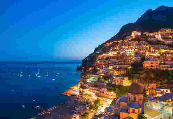 A stunning evening view of the Amalfi Coast, with hillside buildings illuminated against the deep blue sea and sky, and boats scattered along the shoreline. Amalfi Coast_planning a trip to italy and greece_feature