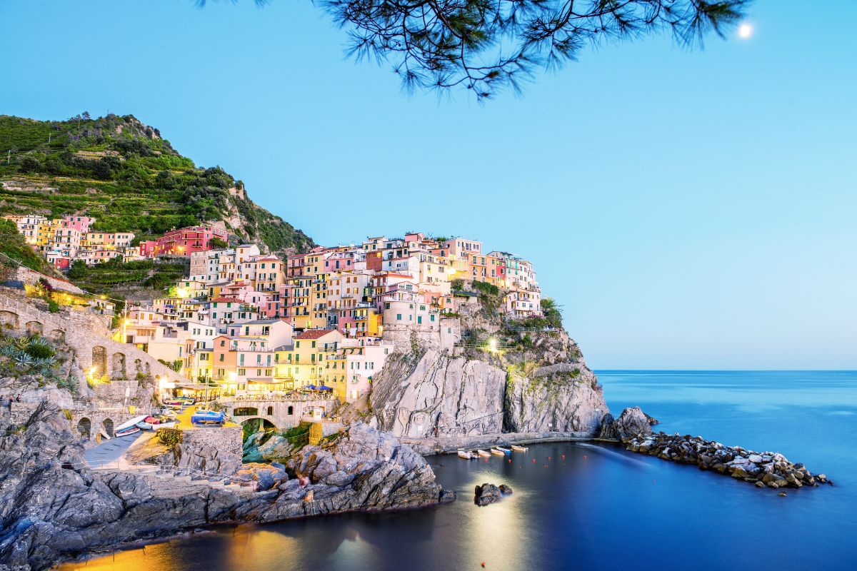 A view of Cinque Terre, featuring colorful hillside houses, rocky cliffs, and a calm sea at dusk.