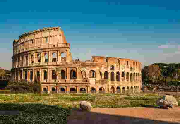 The Colosseum in Rome, bathed in warm sunlight, standing against a clear blue sky with a green field in the foreground. Colloseum_Rome