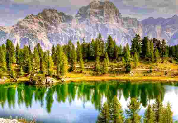 A lake in the Dolomites, Italy, surrounded by green trees with mountains in the background. The water reflects the trees and sky. Dolomites_Weather in Italy and Greece in October_feature