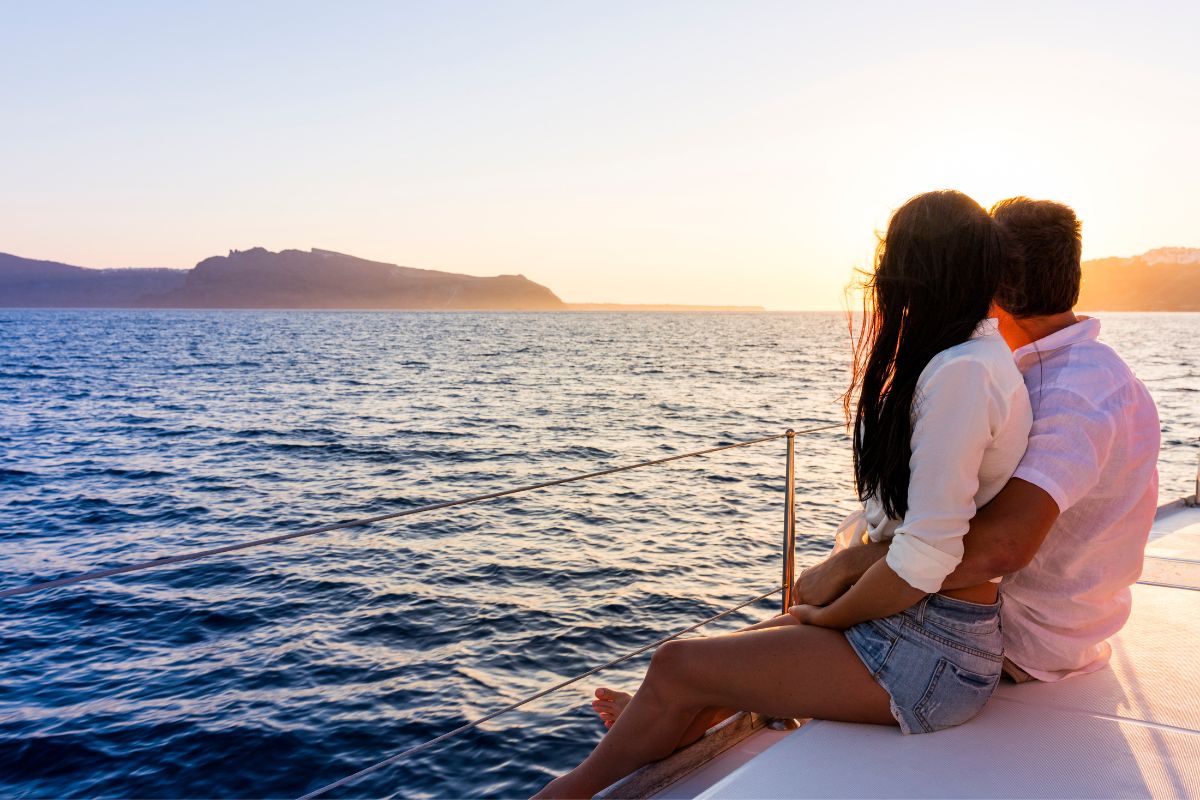 A couple sitting on a yacht, embracing as they watch the sunset over the sea during their honeymoon in Greece.