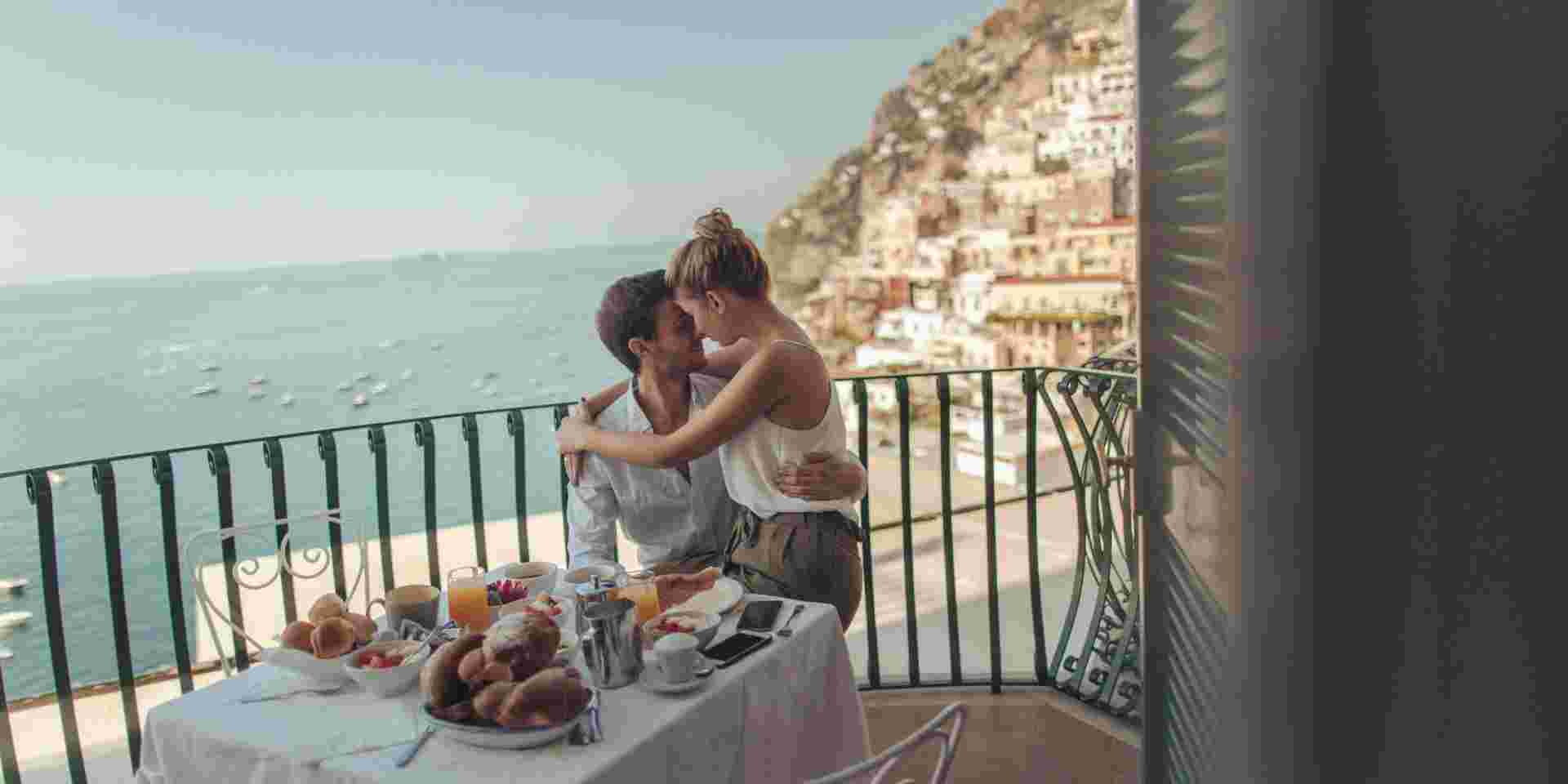 A couple enjoying breakfast on a balcony overlooking the sea and a hillside town in Italy during their honeymoon. Italy and Greece honeymoon_feature