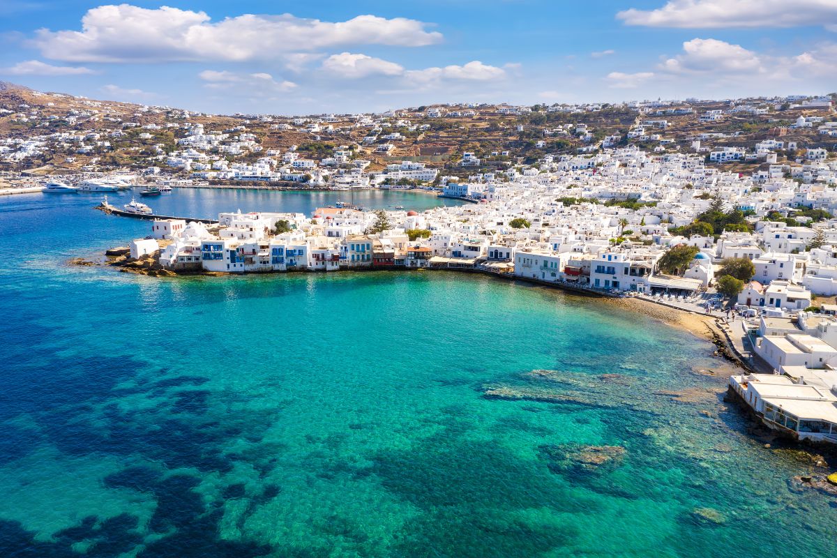 A view of Mykonos, Greece, showing white buildings along the coastline, clear blue waters, and hilly terrain in the background.