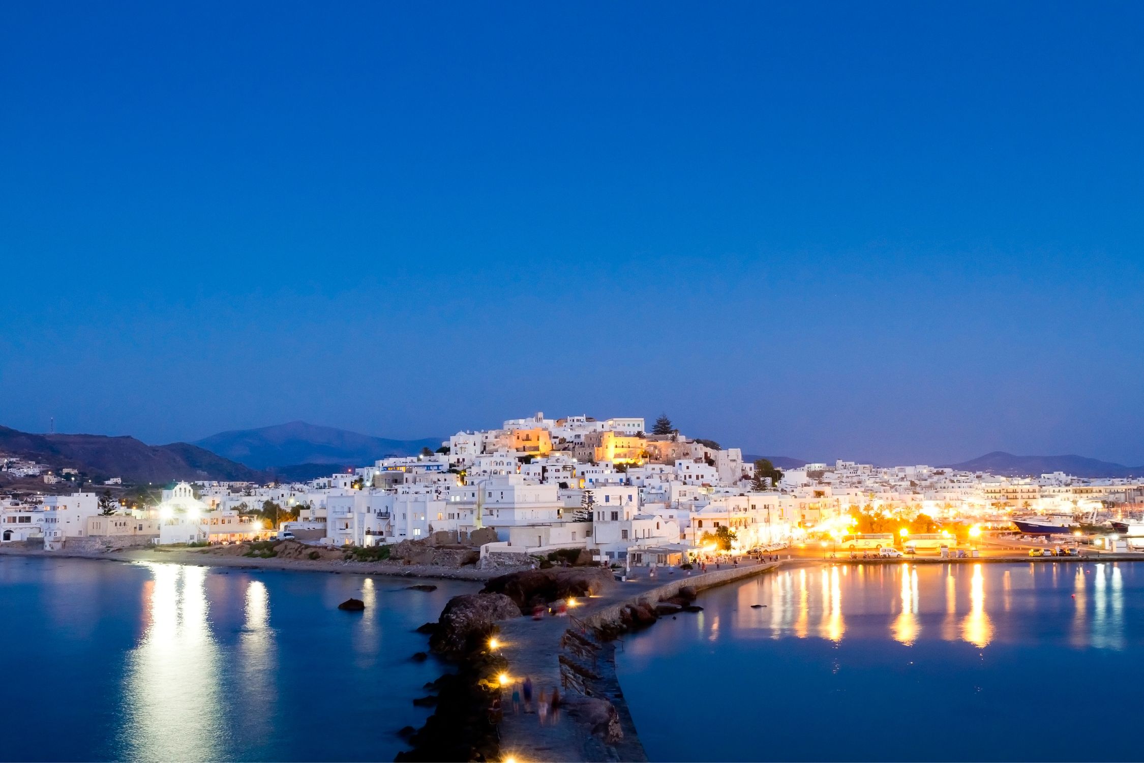 A stunning evening view of Naxos Island, Greece, with white-washed buildings glowing under the deep blue sky, reflecting lights onto the calm waters.