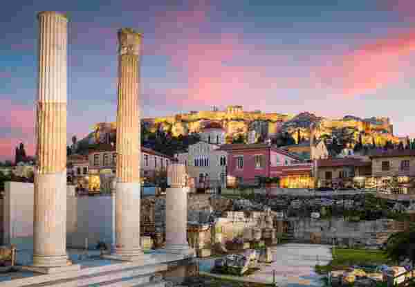A scenic view of Plaka in Athens, Greece, with ancient ruins in the foreground and the illuminated Acropolis atop the hill at sunset. Plaka neighborhood athens