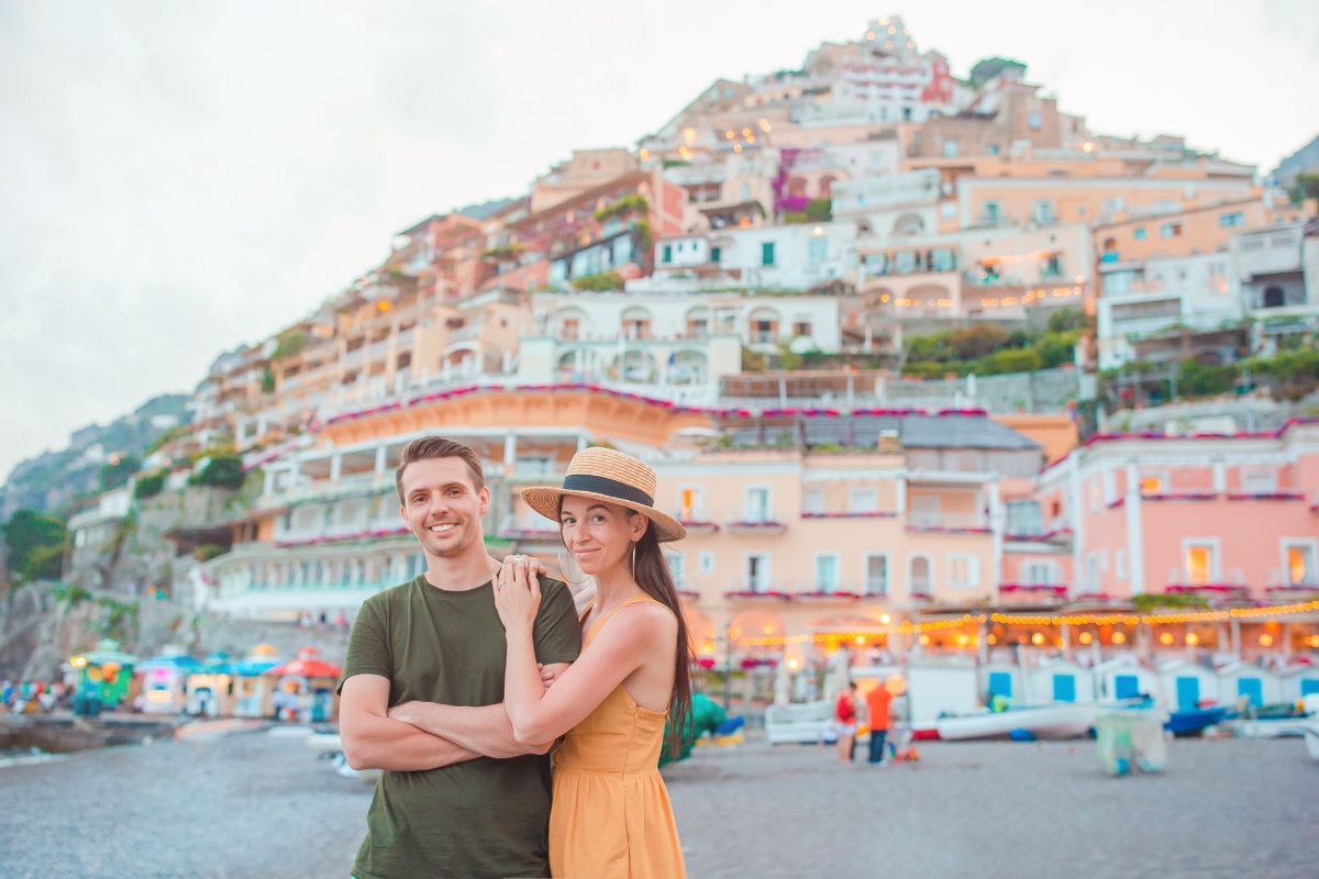 A couple posing on the beach in Positano during their honeymoon, with colorful hillside buildings and lights in the background.