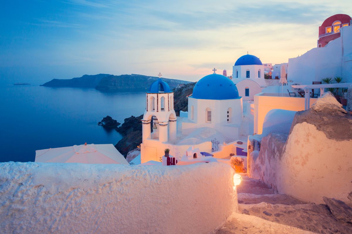 A view of Santorini at dusk, featuring white buildings with blue domes overlooking the calm sea.