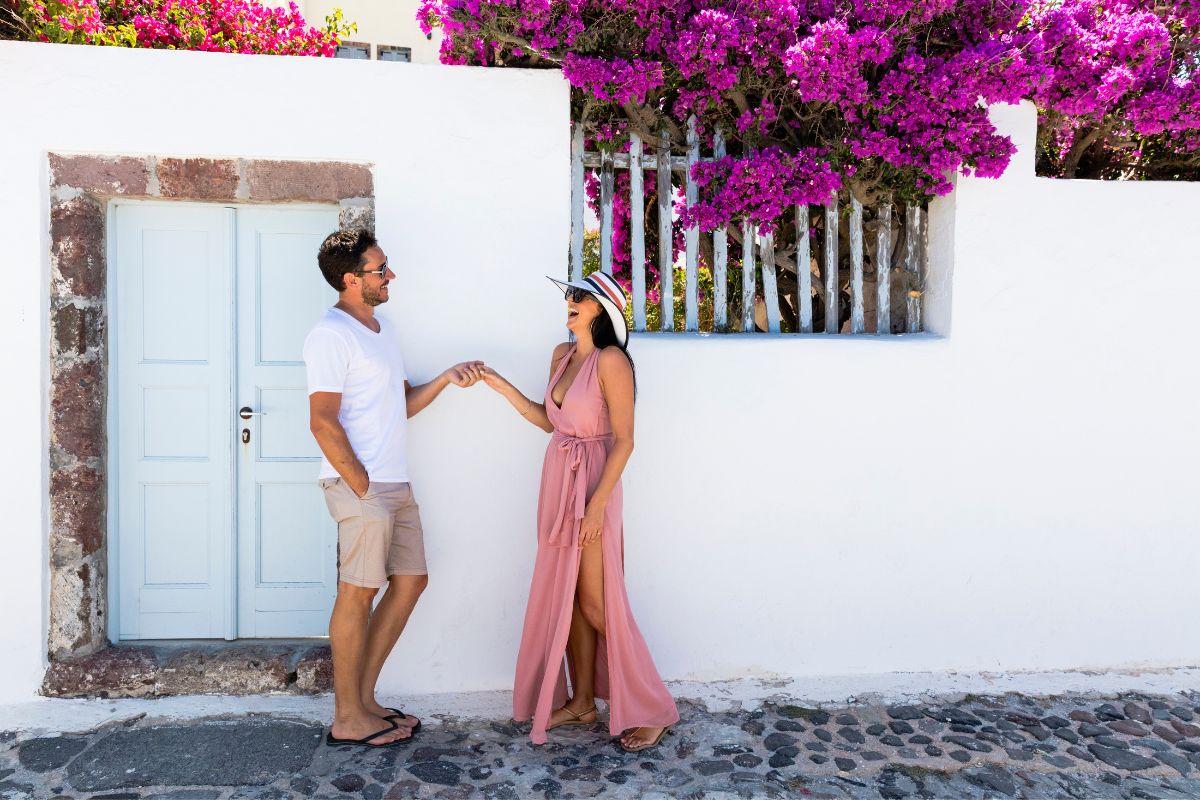 A couple holding hands and smiling in front of a white wall with vibrant pink bougainvillea in Santorini during their honeymoon.