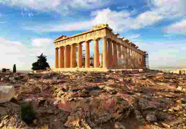 A view of the Parthenon at the Acropolis in Athens, Greece, with its ancient columns and rocky foreground under a partly cloudy sky. acropolis in athens