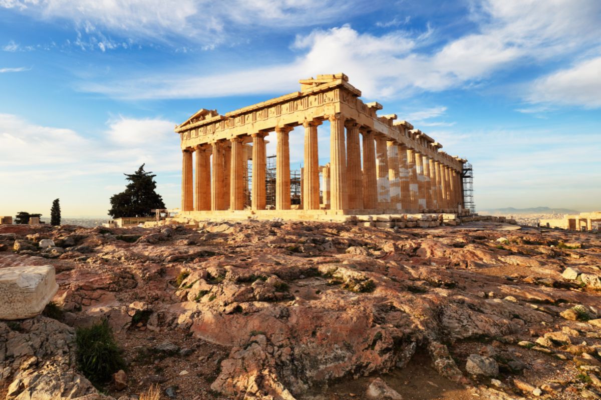 A view of the Parthenon at the Acropolis in Athens, Greece, with its ancient columns and rocky foreground under a partly cloudy sky. acropolis in athens