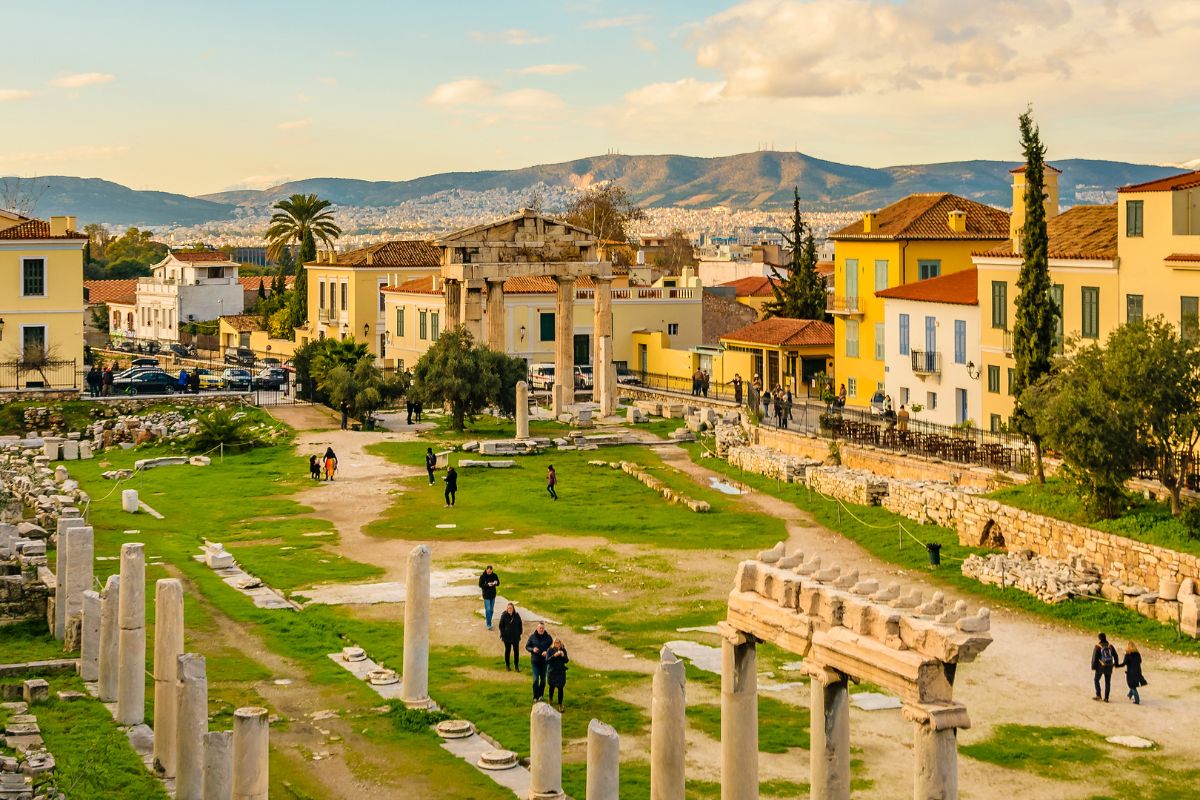 The Ancient Agora of Athens with ruins, scattered columns, and lush greenery, surrounded by colorful buildings and a backdrop of mountains_Italy and Greece 14 Days of Adventure