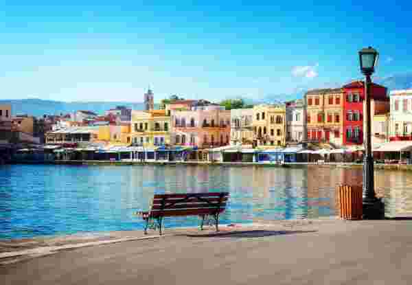 A view of the Chania waterfront in Crete, Greece, featuring colorful buildings, a calm harbor, and a bench overlooking the water. chania crete