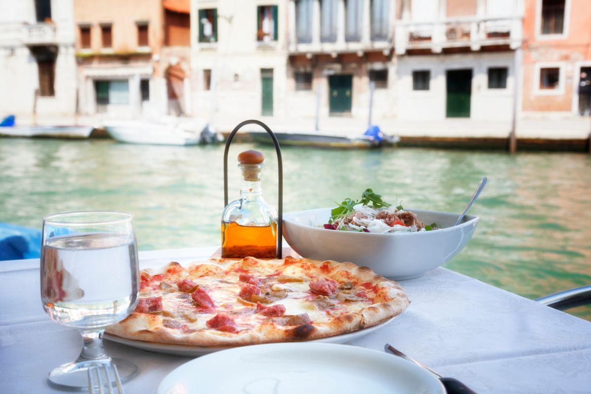 A delicious Italian meal with pizza, salad, and a bottle of olive oil set on a table by a canal, with historic buildings in the background_Planning a Trip to Italy and Greece
