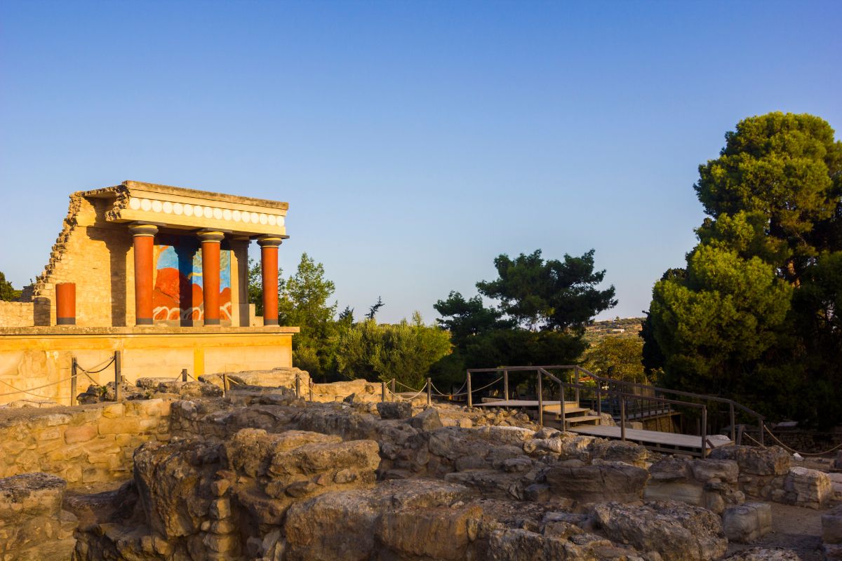 The ruins of Knossos Palace in Crete, Greece, with red columns and a restored fresco. Stone structures and trees surround the site under a clear sky.