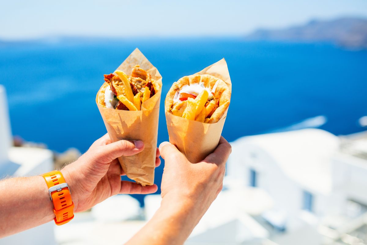 Two hands holding pita gyros wrapped in paper, filled with meat, fries, and toppings, with a blue sea and white buildings in the background.