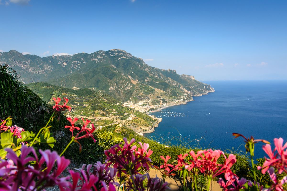 A breathtaking view of Ravello, Italy, with colorful flowers in the foreground, lush green mountains, and the blue Mediterranean Sea stretching into the horizon.
