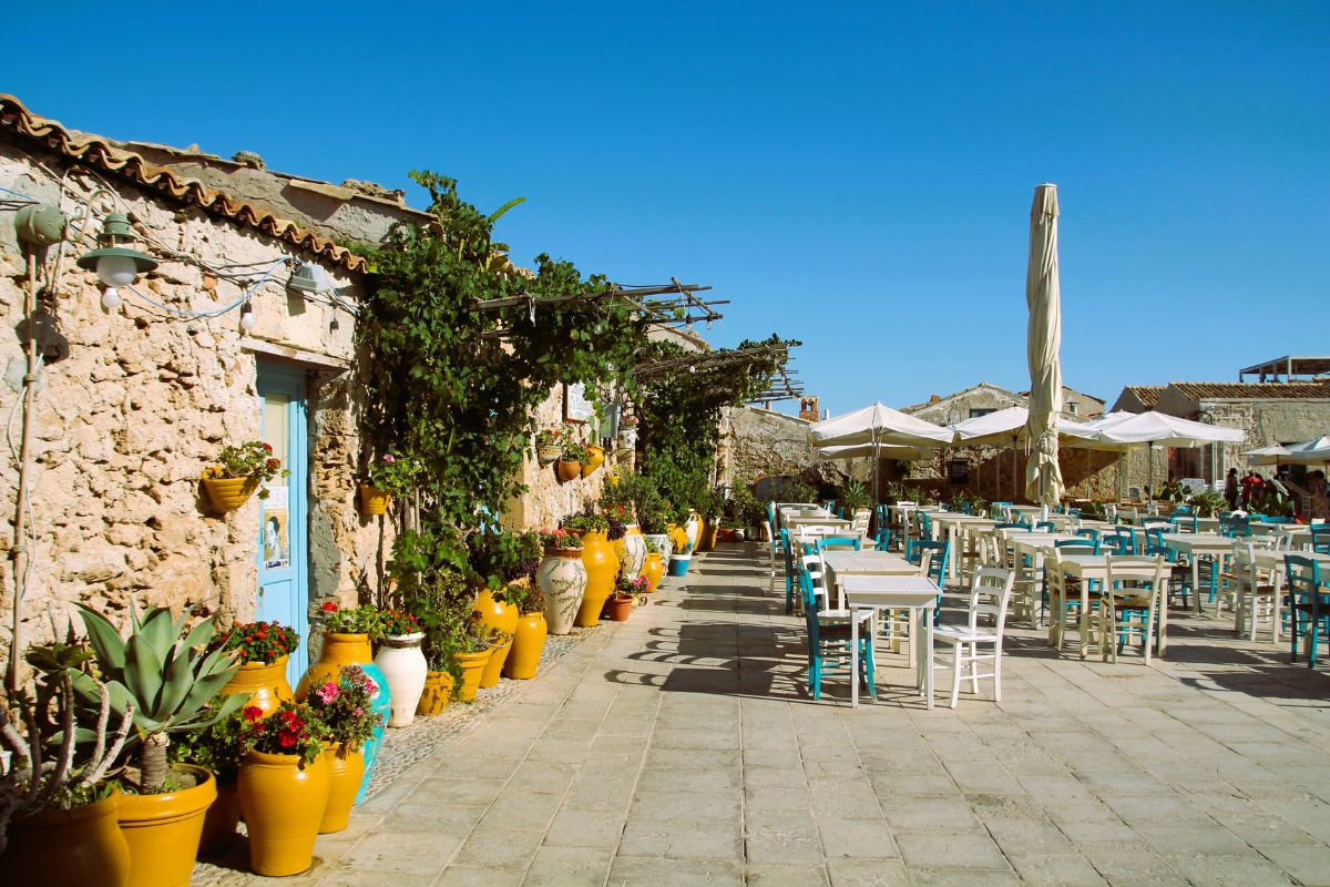 Outdoor café with colorful pots and stone walls in Sicily, showcasing sunny Mediterranean charm in October.