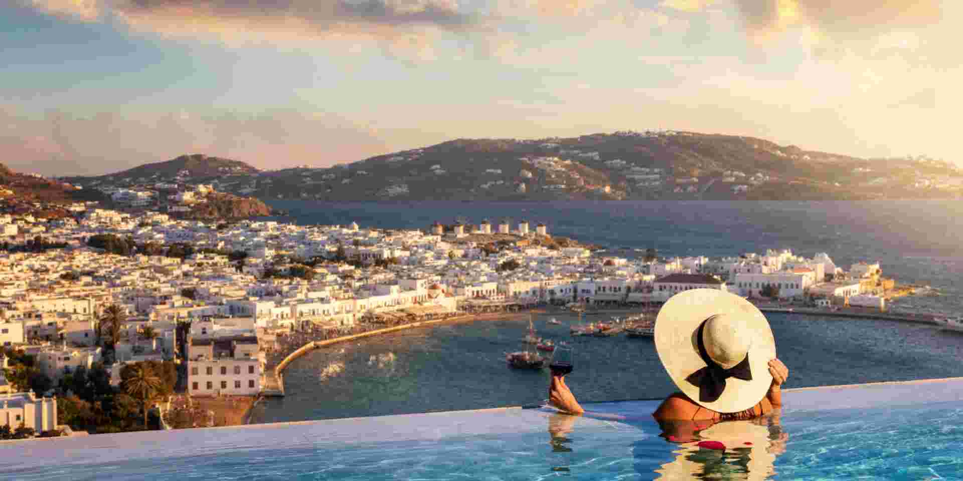 A tourist woman with a glas of wine in a swimming pool enjoys the view over the town of Mykonos island, Greece, during summer sunset time Mykonos hotels_feature