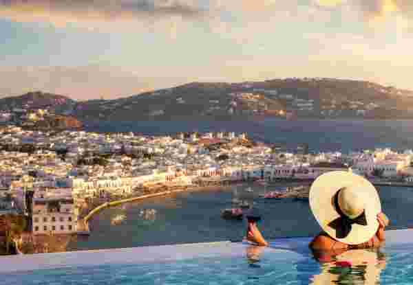 A tourist woman with a glas of wine in a swimming pool enjoys the view over the town of Mykonos island, Greece, during summer sunset time Mykonos hotels_feature