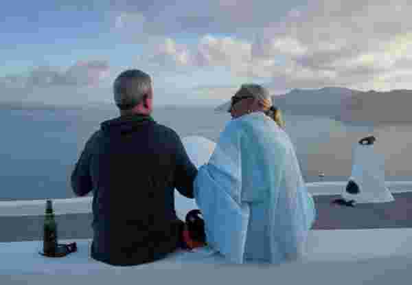 Senior couple travelers enjoy Santorini summer vacation sitting on wall having a beer drink and looking at amazing panoramic view over the roofs santorini for seniors_feature