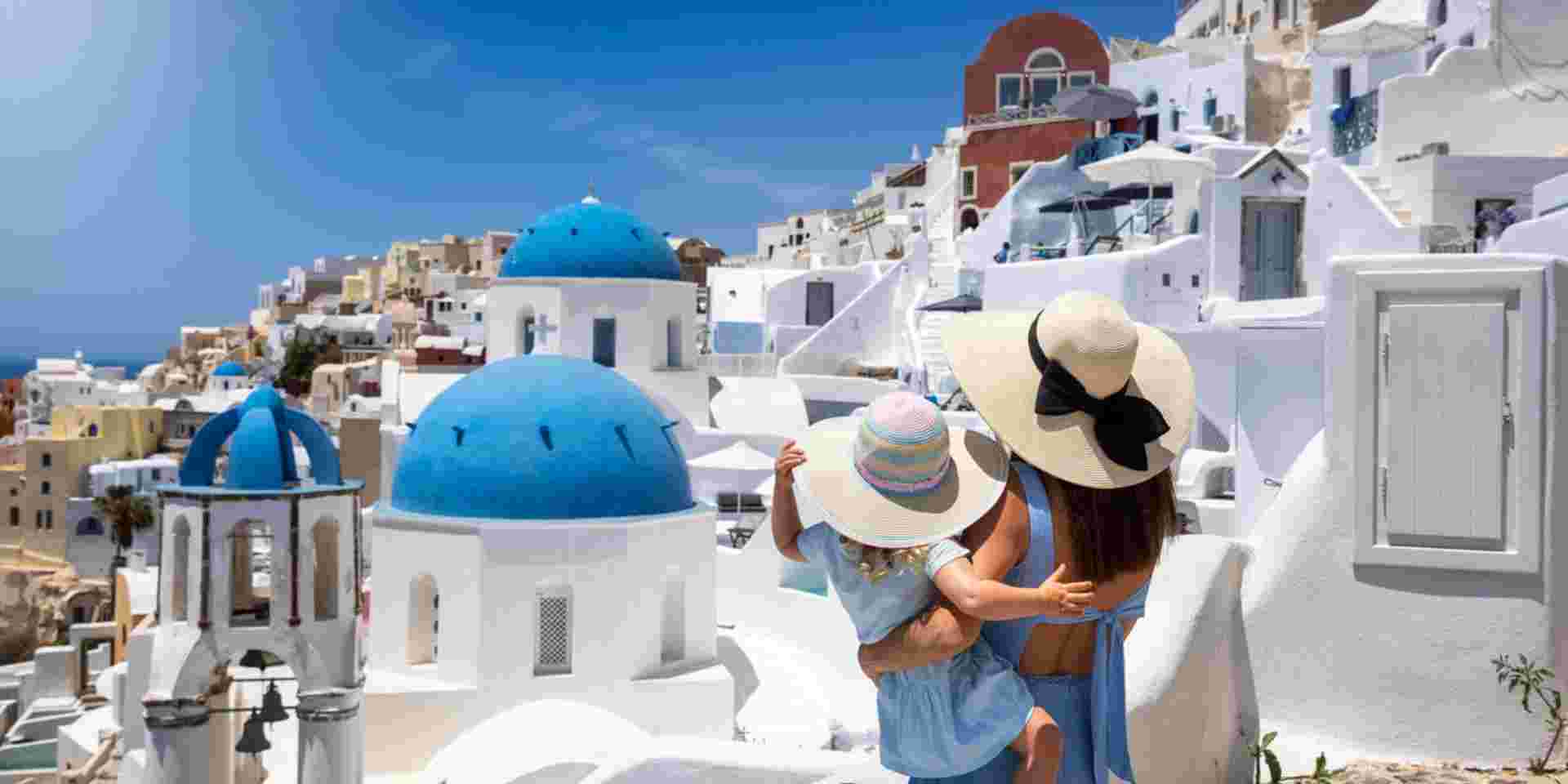 A tourist mother and her daughter looking at the whitewashed houses of the village Oia, Santorini island, Greece, during summer holiday time santorini with kids_feature