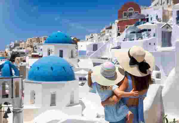 A tourist mother and her daughter looking at the whitewashed houses of the village Oia, Santorini island, Greece, during summer holiday time santorini with kids_feature