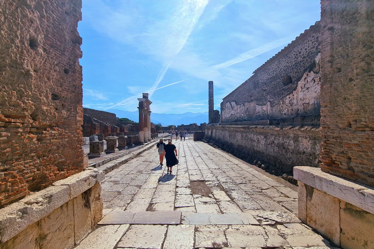 Visitors walking along an ancient stone street in the ruins of Pompeii, Italy, under a clear blue sky.