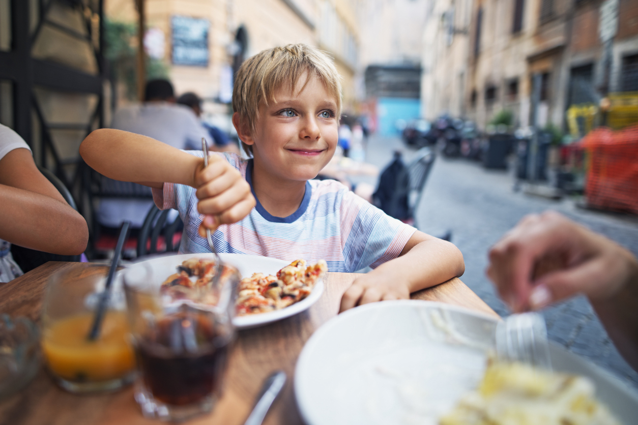 Little boy aged 7 having lunch with family in street restaurant in Rome, Italy. The girl is smiling and and looking sideways while getting a piece of pizza on the fork. Family is enjoying a happy meal together.