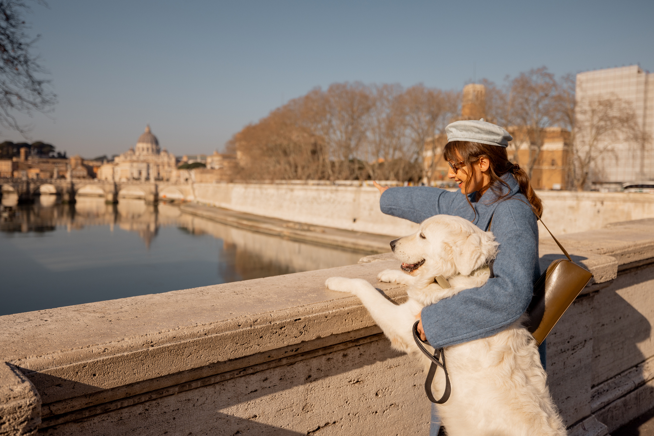 A woman in a blue coat embraces her large white dog on a Roman bridge, gazing toward St. Peters Basilica under a clear winter sky