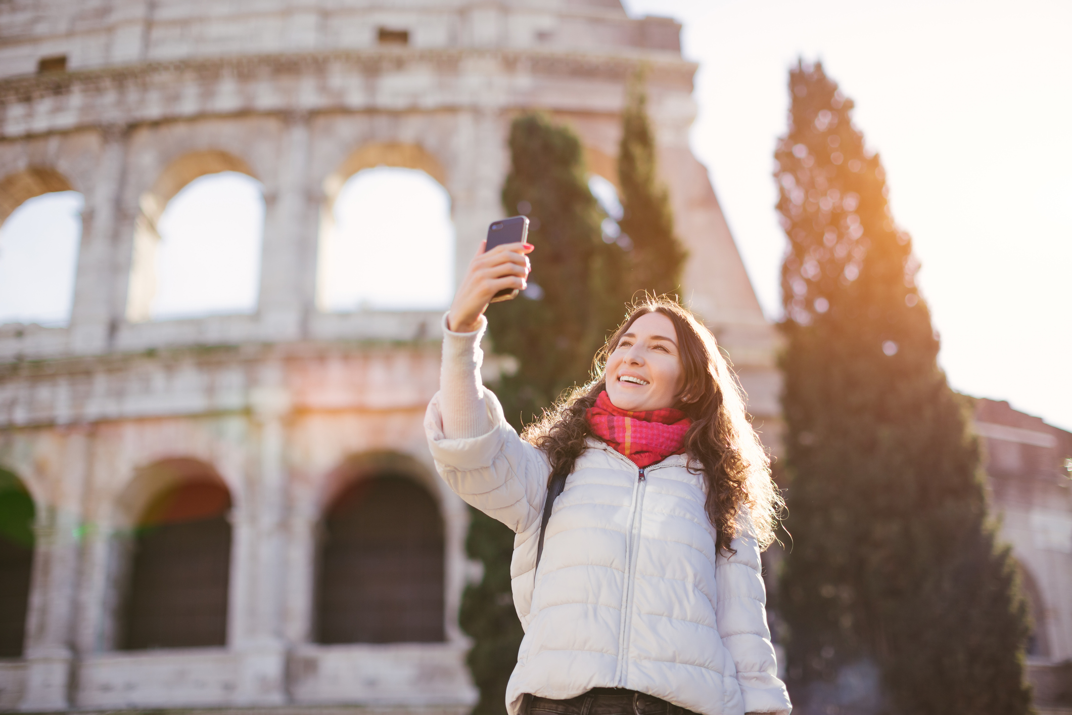 Young beautiful smiling woman posing against Roman colosseum (Coliseum) and making selfie with smartphone - winter holidays and vacation in Europe concept