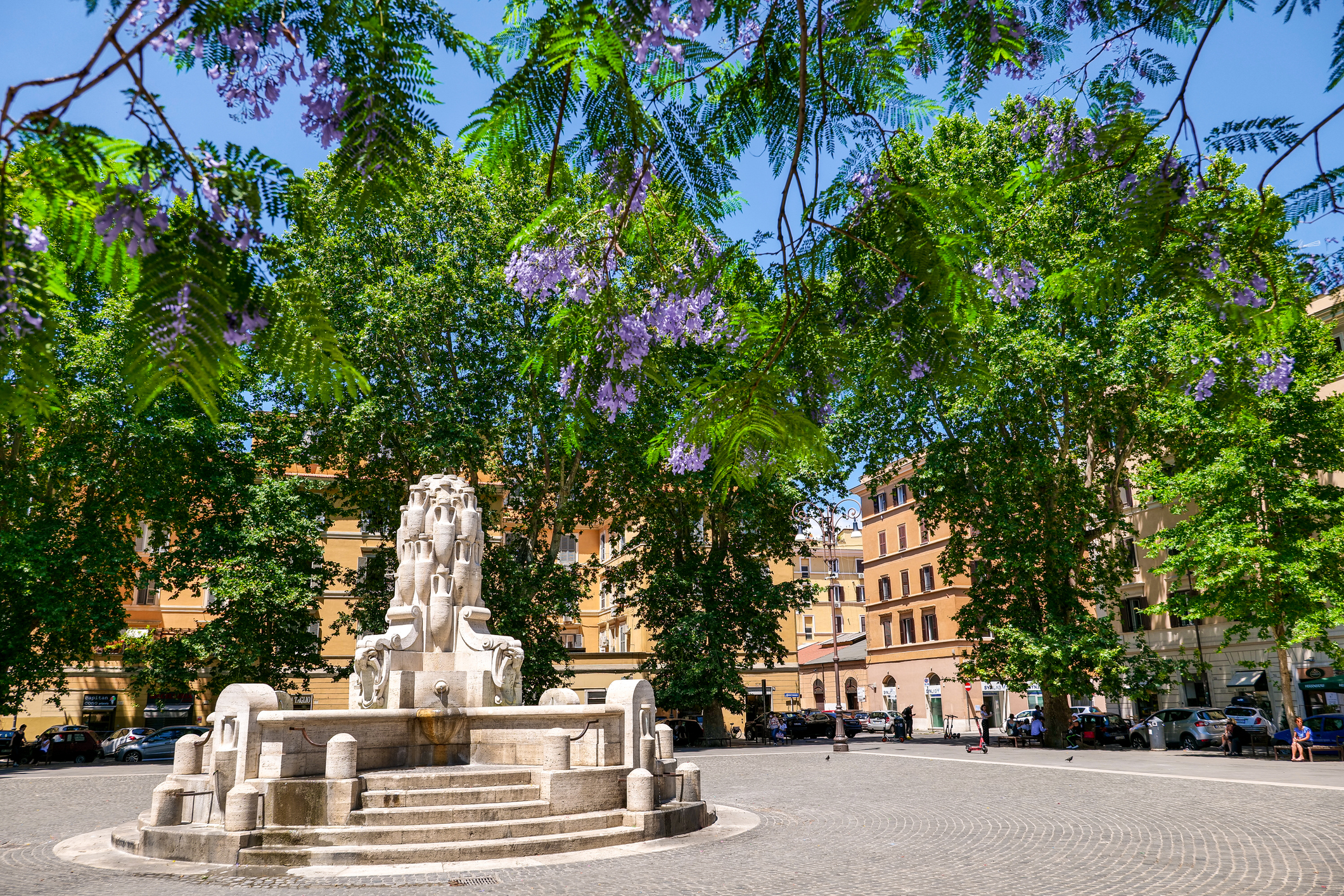 Rome, Italy, June 15 -- A sunny view of the Testaccio Square, near the Ostiense district, with the Fontana delle Anfore (Fountain of the Amphorae), made in 1927 by the sculptor Pietro Lombardi, and some flowered jacaranda tree. Present in the topography of Rome since the imperial era due to the presence of the river port on the Tiber, Testaccio is one of the oldest districts of the Italian capital, visited by tourists for the presence of numerous museums, cultural and youth centers, typical Roman restaurants, remains from Roman times and experimental musical and theatrical spaces. Testaccio maintains a strong popular identity, where to find the true soul of Rome. Image in High Definition format.