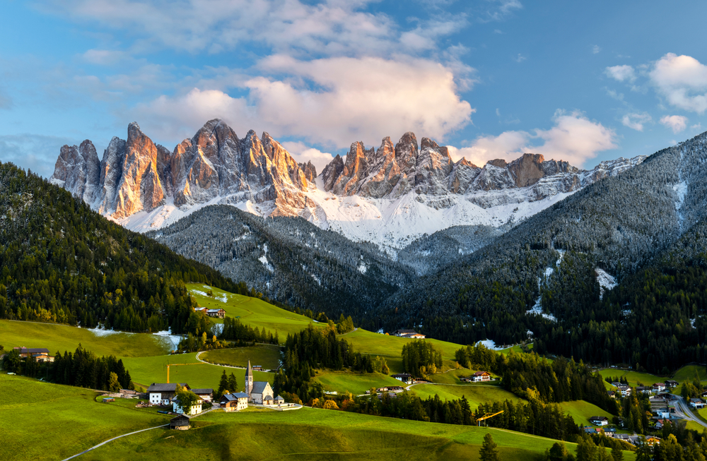 Panoramic view of the Dolomites with snow-capped peaks, rolling green meadows and a traditional alpine village at sunset