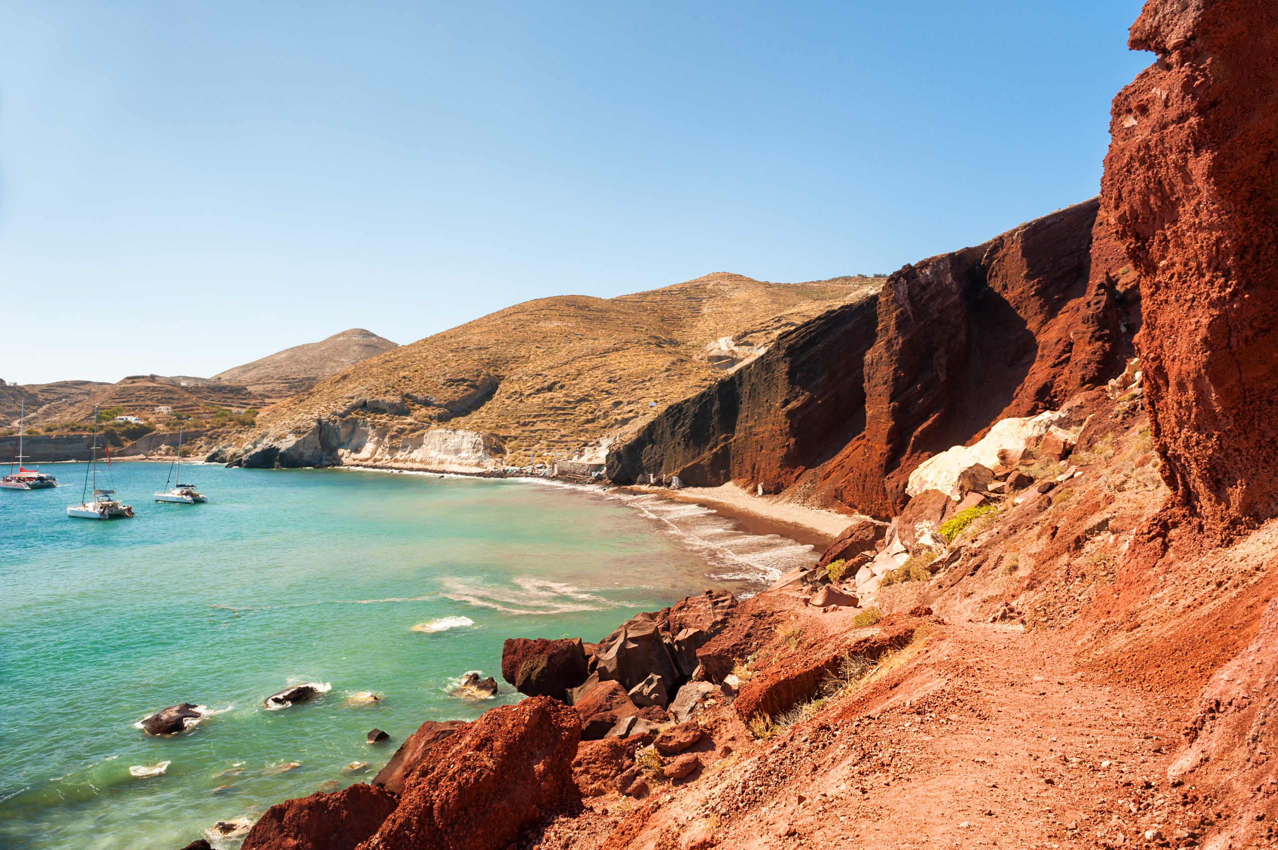 Sailboats anchored in a turquoise cove beneath Santorini's Red Beach cliffs.