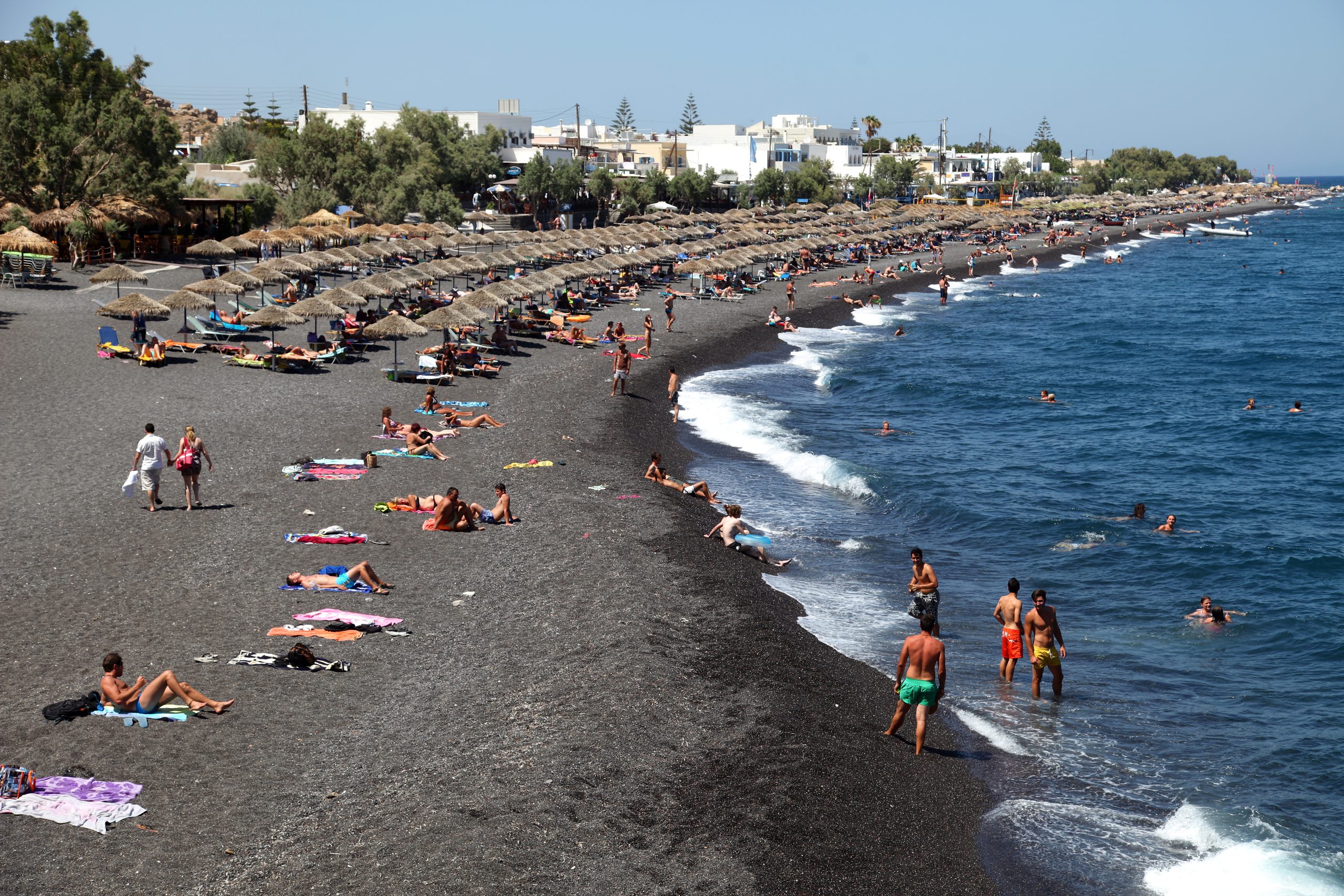 Rows of straw umbrellas and sunbathers on Kamari's black pebble beach beside the sea. 