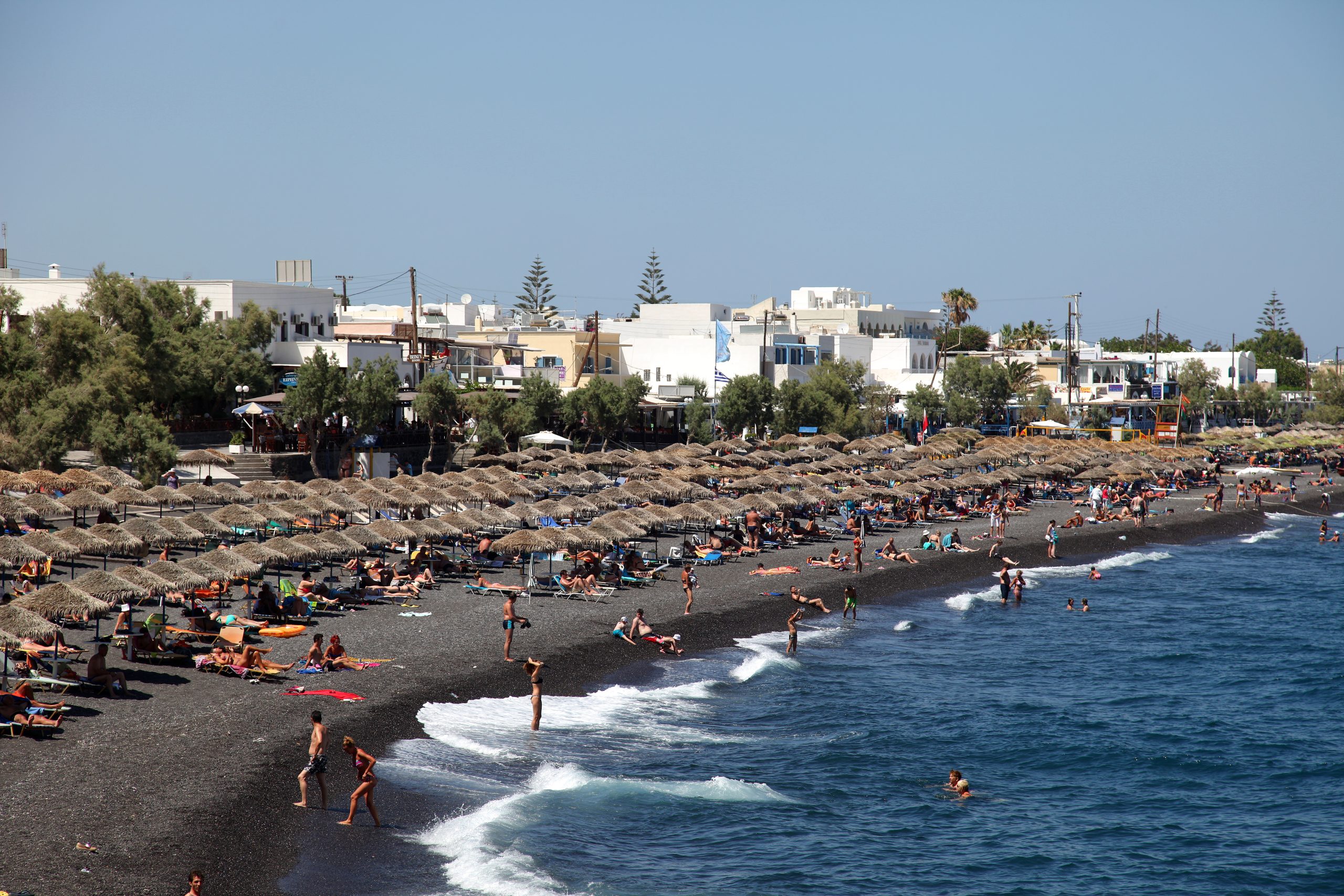 Sunbathers and straw umbrellas lining Kamari's black sand beach, with whitewashed buildings behind.