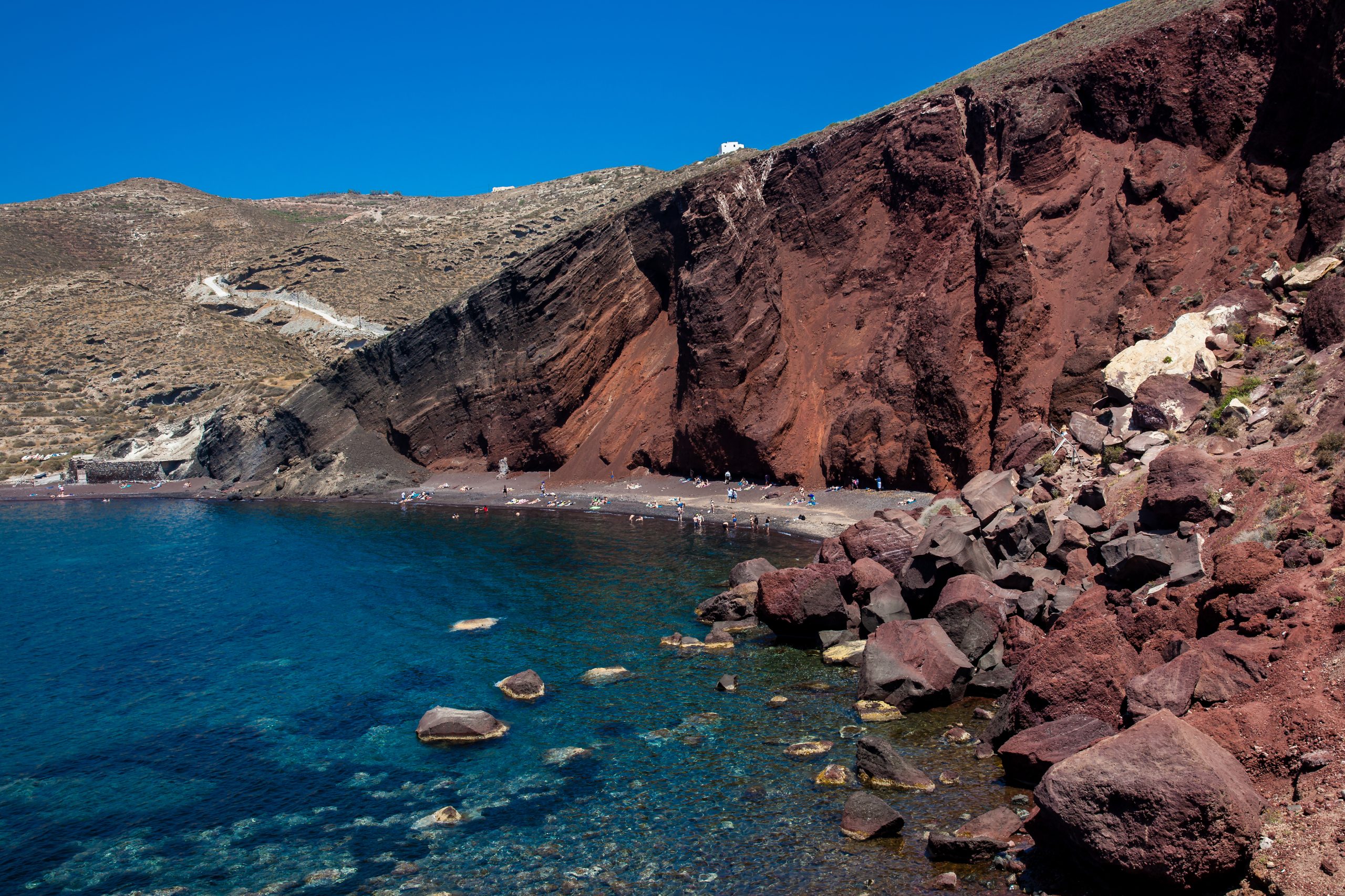 Sunbathers on Santorini's Red Beach, tucked beneath towering red volcanic cliffs beside clear blue water.