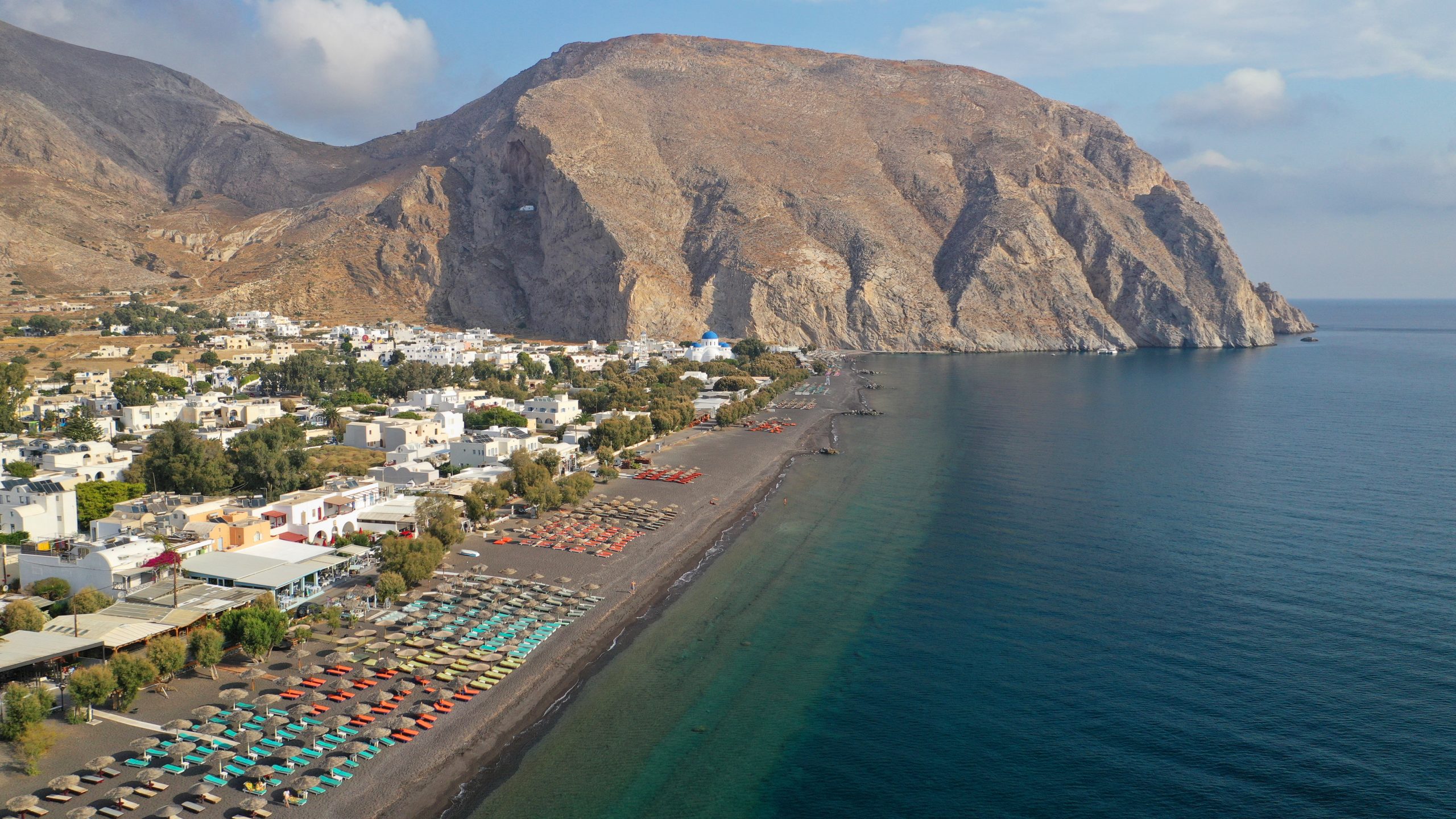 Aerial view of Perissa Beach's long black sand shoreline, lined with umbrellas beneath towering cliffs.