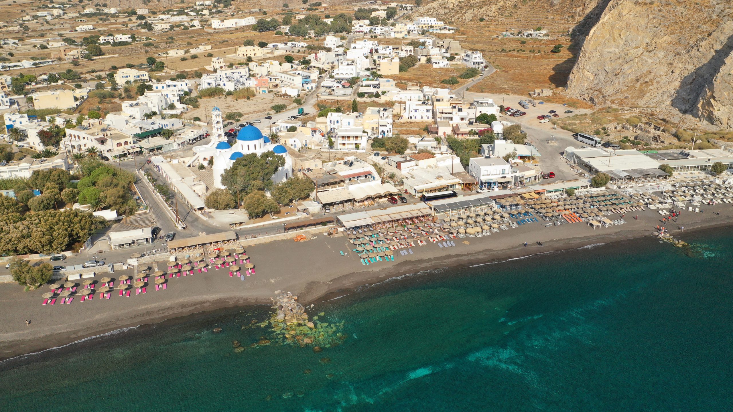 Aerial view of Perissa's black sand beach, blue-domed church, and whitewashed village beneath a rocky Santorini cliff. 