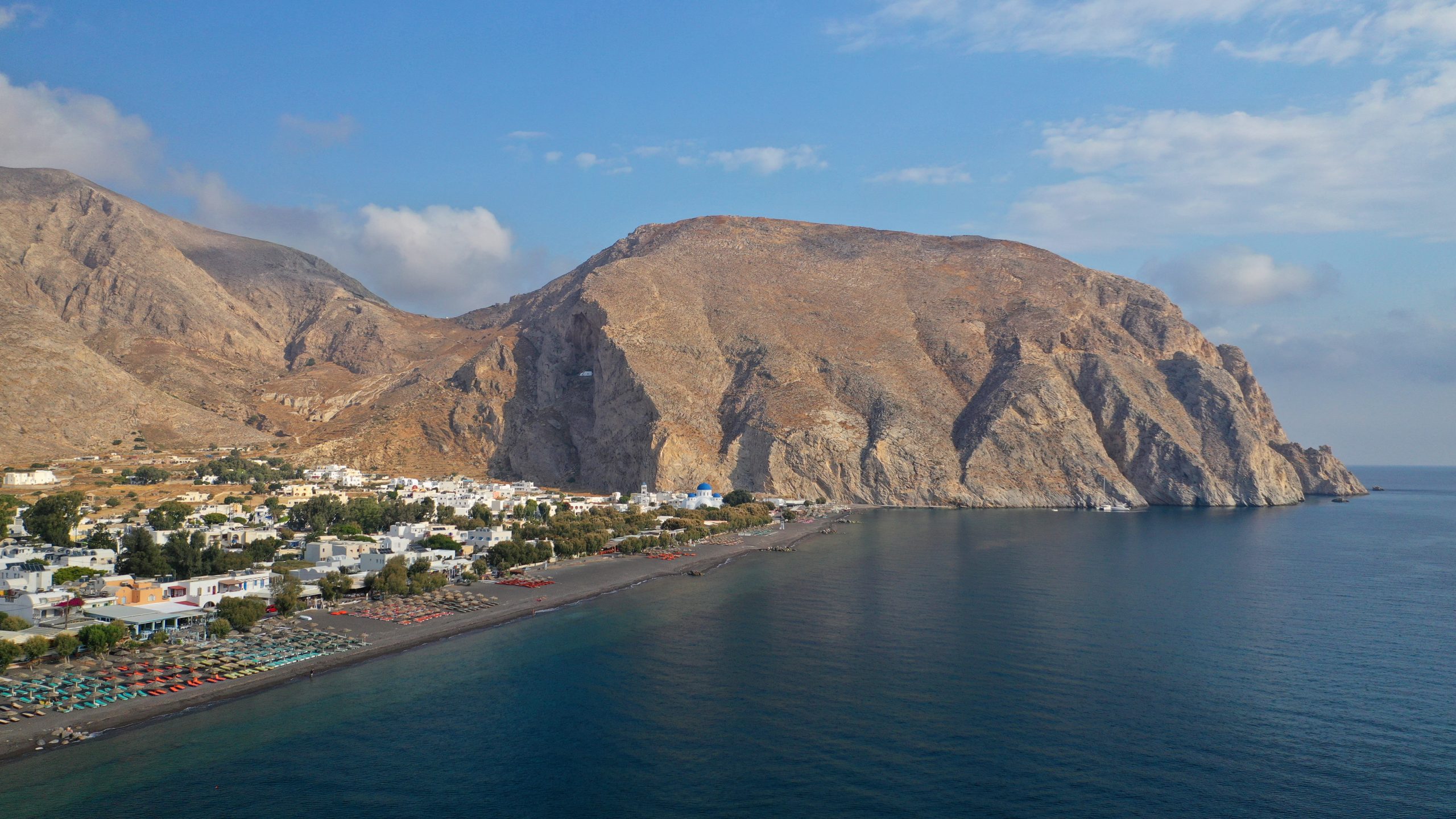 Aerial view of Kamari's dark sand beach curving beneath rugged cliffs on Santorini's coast.