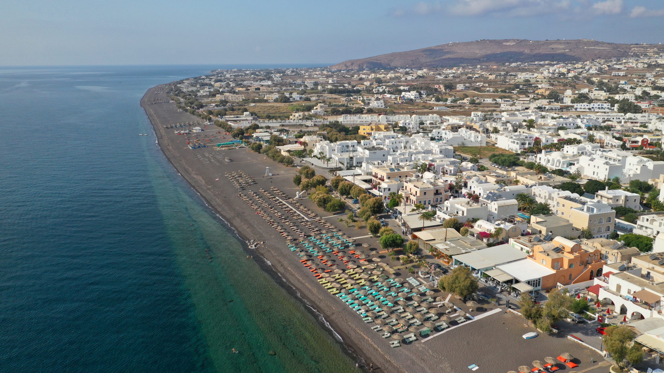 Aerial view of Perissa's black sand beach and whitewashed village stretching along Santorini's southeastern coast. 