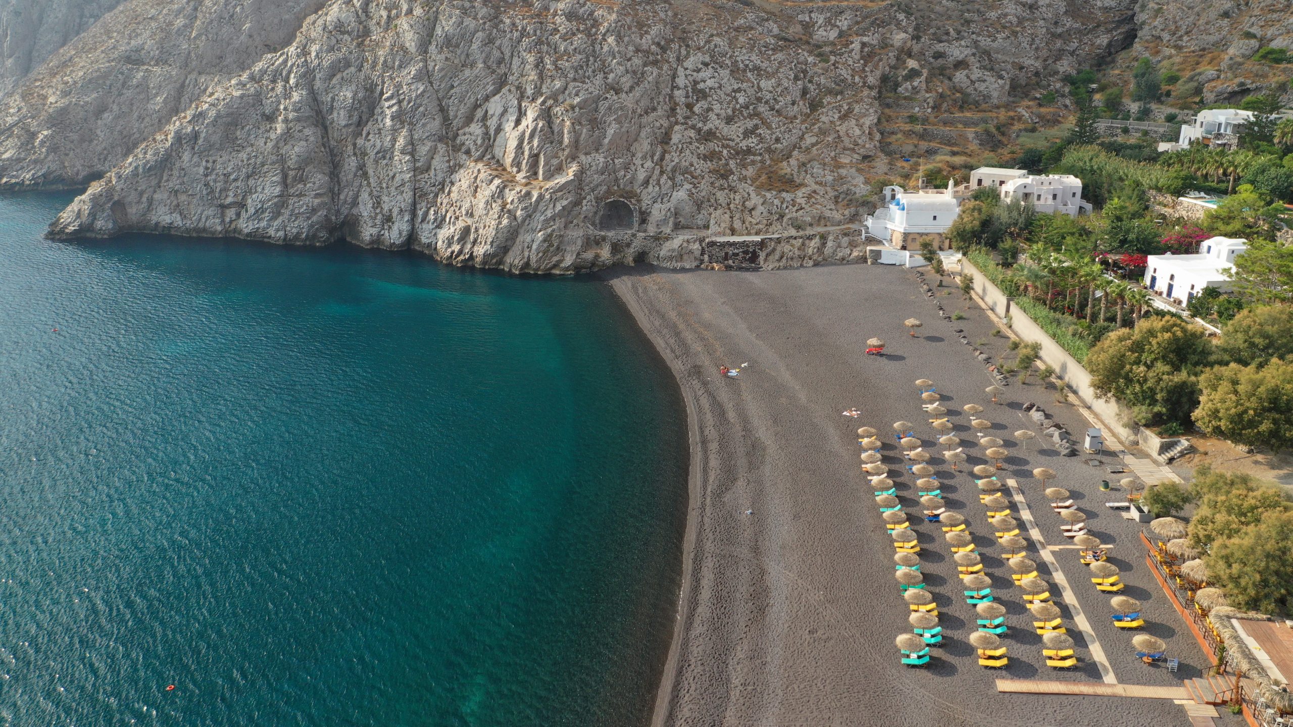 Aerial view of Perissa Beach, Santorini, with colorful sun loungers lining the black pebble shore beside a whitewashed church.