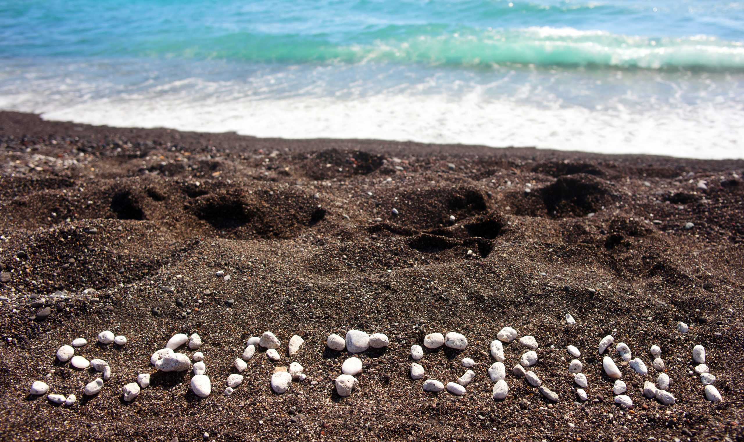 Close-up of white pebbles arranged on black volcanic sand beside gentle turquoise waves. 