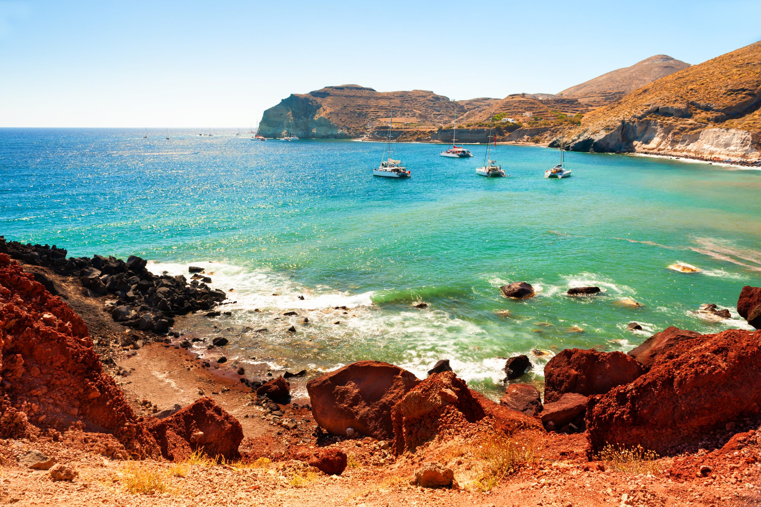 Sailboats anchored in a turquoise cove near Santorini's red volcanic cliffs.