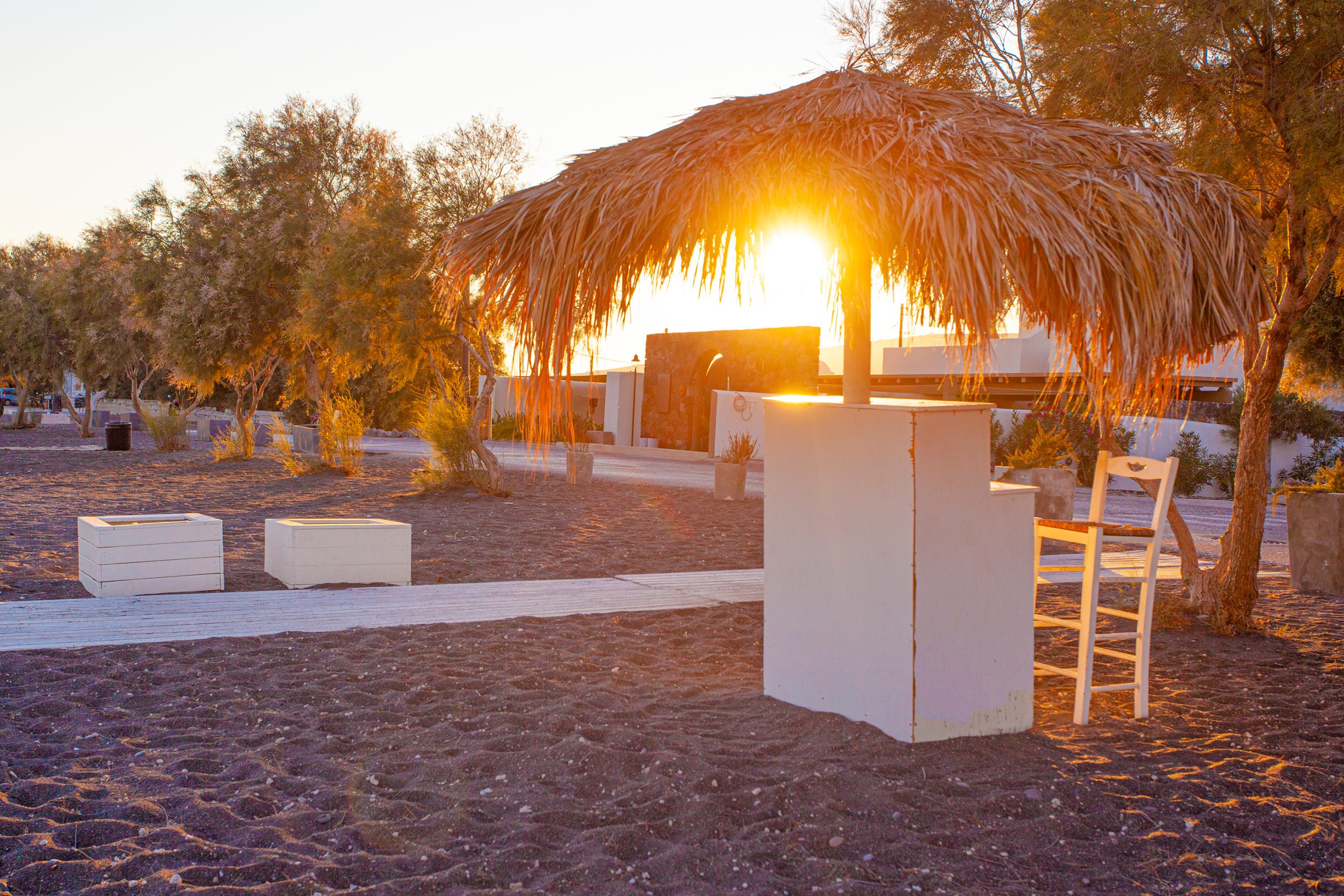 Golden sunset light filtering through a straw beach umbrella on Santorini's black sand shore. 