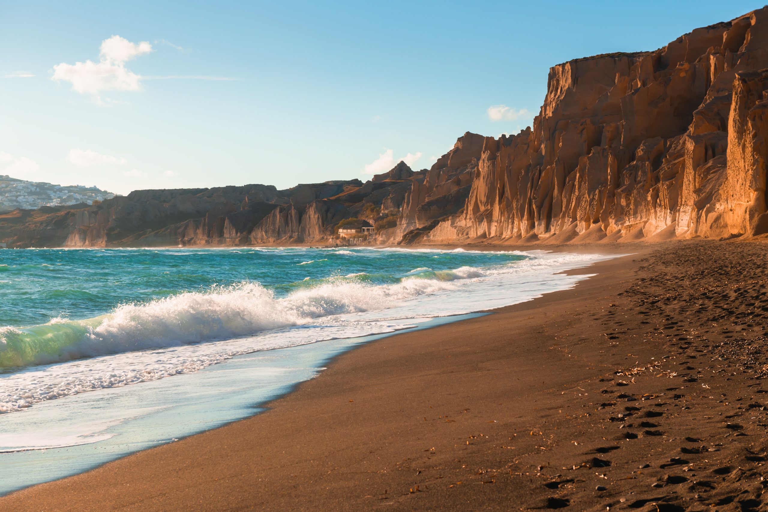 Waves crashing on Vlychada's black sand beach beneath dramatic Santorini cliffs at golden hour.