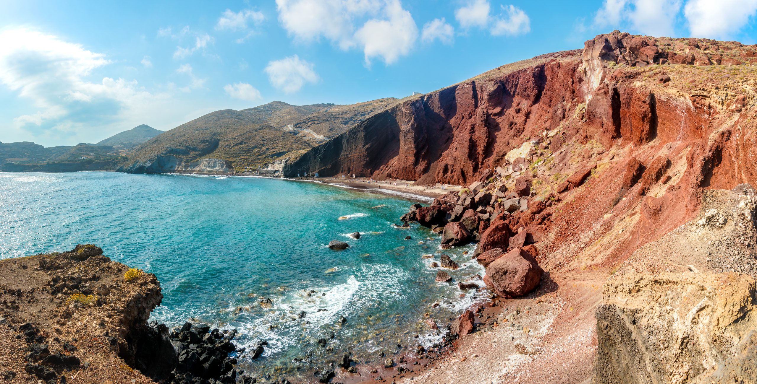 Panoramic view of Red Beach, Santorini, with dramatic rust-colored volcanic cliffs above turquoise water.